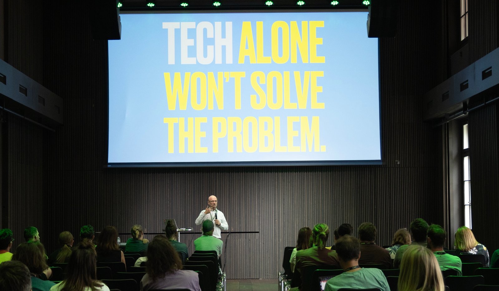 Christian Hansen als Speaker vor einem grossen Publikum in einem Saal. Hinter ihm eine sehr grosse Leinwand, auf die in sehr grossen Lettern der Satz "Tech alone won't solve the problem" projiziert ist.