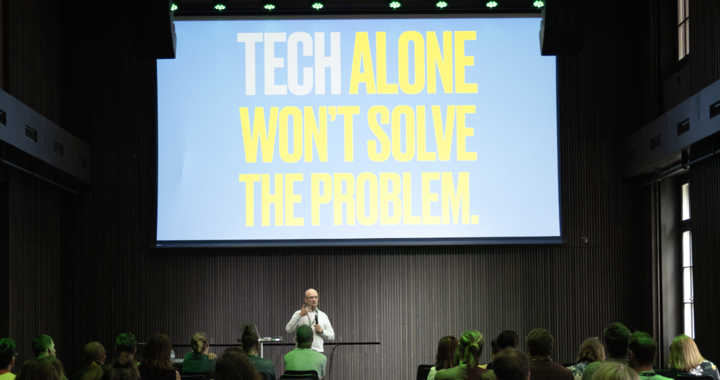 Christian Hansen steht als Referent vor einem grösseren Publikum und spricht in ein Mikrofon. Hinter ihm wird auf eine sehr grosse Leinwand der in bunten, sehr fetten Lettern der Satz projiziert: "Tech alone won't solve the problem". 