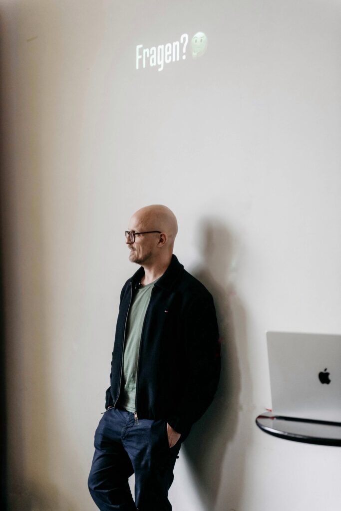 Christian Hansen bei einem Auftritt. Er lehnt an einer Wand, schaut ins Publikum. Neben ihm ein Laptop, den er für seine Präsentation genutzt hat. Über ihm projiziert an die Wand das Wort "Fragen?"